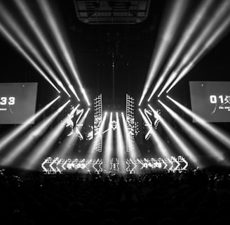 A black and white image captures a dramatic indoor scene featuring a large stage setup with intense beams of light radiating from the center. Two large screens are prominently displaying the text '01:33 ESL One Cologne 2017'. The audience can be seen seated in the dark foreground, while angular patterns can be spotted on the stage.