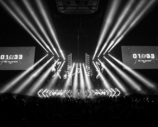 A black and white image captures a dramatic indoor scene featuring a large stage setup with intense beams of light radiating from the center. Two large screens are prominently displaying the text '01:33 ESL One Cologne 2017'. The audience can be seen seated in the dark foreground, while angular patterns can be spotted on the stage.