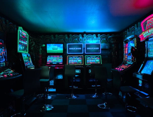 Dimly lit gaming arcade featuring a variety of brightly illuminated slot machines and video gaming screens. Neon signs and colorful displays create a vibrant atmosphere. The arrangement of gaming machines suggests an inviting space for entertainment, with chairs positioned for players.