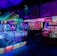 An indoor arcade filled with vibrant gaming machines lit with neon blue and pink lights. The scene includes various arcade games with flashing screens and colorful displays, and a large overhead structure resembling a green snake. The background shows illuminated signs and themed decor, contributing to an immersive entertainment atmosphere.