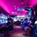 A vibrant arcade with rows of gaming machines adorned with colorful screens. Several people are immersed in playing games, seated on stools in a dimly lit room accented by an intense pink ceiling light. The atmosphere is lively and engaging.