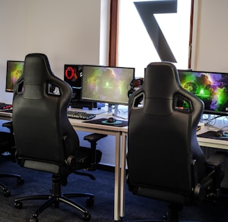 A row of gaming stations equipped with large monitors, gaming chairs, keyboards, and headphones. The setup is organized and ready for gaming, featuring a colorful screen display. The room has a modern and tech-savvy ambiance.
