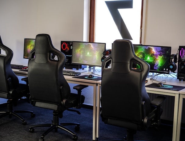 A row of gaming stations equipped with large monitors, gaming chairs, keyboards, and headphones. The setup is organized and ready for gaming, featuring a colorful screen display. The room has a modern and tech-savvy ambiance.