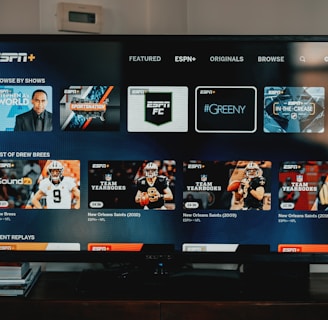 A television screen displaying the ESPN+ streaming interface with various sports-related shows and categories. The screen includes sections such as 'Browse by Shows' and 'Best of Drew Brees' with thumbnails of sports programs featuring athletes in action and analysts.