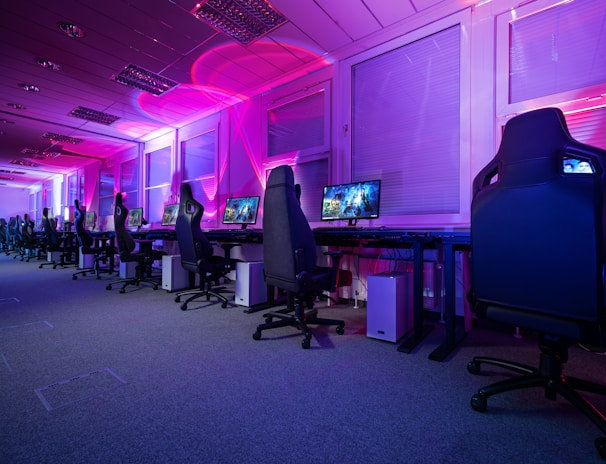 A row of gaming computers with large monitors and racing-style chairs is set up along the interior of a modern room. The space is illuminated with vibrant purple and blue LED lighting, creating a futuristic atmosphere. The room is organized and clean, suggesting a professional gaming or esports environment.