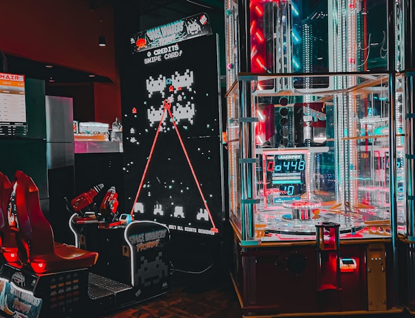 An arcade setting featuring a classic Space Invaders game setup and a modern gaming machine with seats. Bright neon lights illuminate the area, with scores displayed on digital screens. A bright and visually engaging environment full of electronic components.