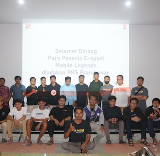 A group of people is posing for a photograph in a room with a white screen in the background that has text welcoming e-sport participants for a Mobile Legends event organized by PKS Prambanan. The group consists of 20 individuals standing and sitting, with some wearing casual attire and others wearing matching shirts that include logos.