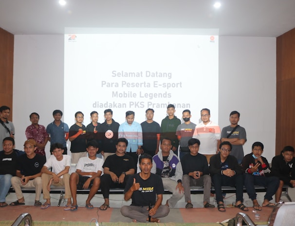 A group of people is posing for a photograph in a room with a white screen in the background that has text welcoming e-sport participants for a Mobile Legends event organized by PKS Prambanan. The group consists of 20 individuals standing and sitting, with some wearing casual attire and others wearing matching shirts that include logos.