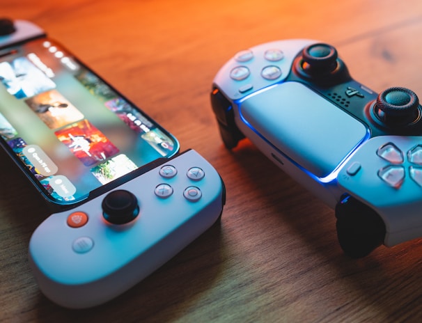 A gaming scenario featuring a smartphone with a game interface next to two controllers on a wooden surface. The smartphone displays vibrant images and icons, while the controllers feature a sleek, modern design with buttons and joysticks prominently visible. Warm lighting enhances the scene.