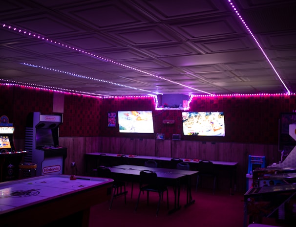 A dimly lit room with vibrant purple LED strip lights on the ceiling, featuring multiple gaming stations. There are two large screens showing video games, along with various arcade machines, including a Nintendo arcade cabinet. An air hockey table is positioned in the foreground.
