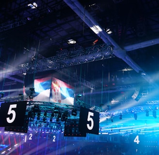 A large indoor arena is equipped with advanced lighting and audio systems. Suspended above the space are numerous digital displays showcasing numbers. The environment is filled with blue illumination and spotlights creating a vibrant atmosphere.