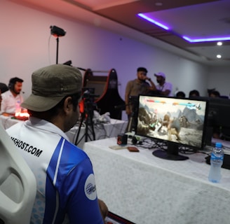 A group of people is gathered in a room watching and participating in a video game competition. Players and spectators are focused on the monitors displaying a fighting game. The setting includes tables with equipment and chairs arranged around the venue, with a noticeable intensity in the participants’ engagement.