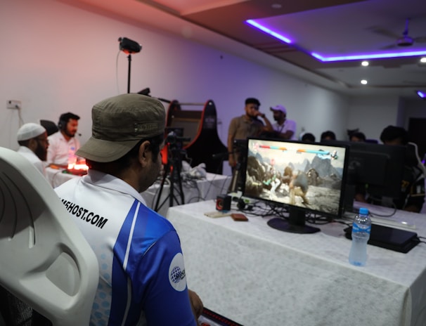 A group of people is gathered in a room watching and participating in a video game competition. Players and spectators are focused on the monitors displaying a fighting game. The setting includes tables with equipment and chairs arranged around the venue, with a noticeable intensity in the participants’ engagement.